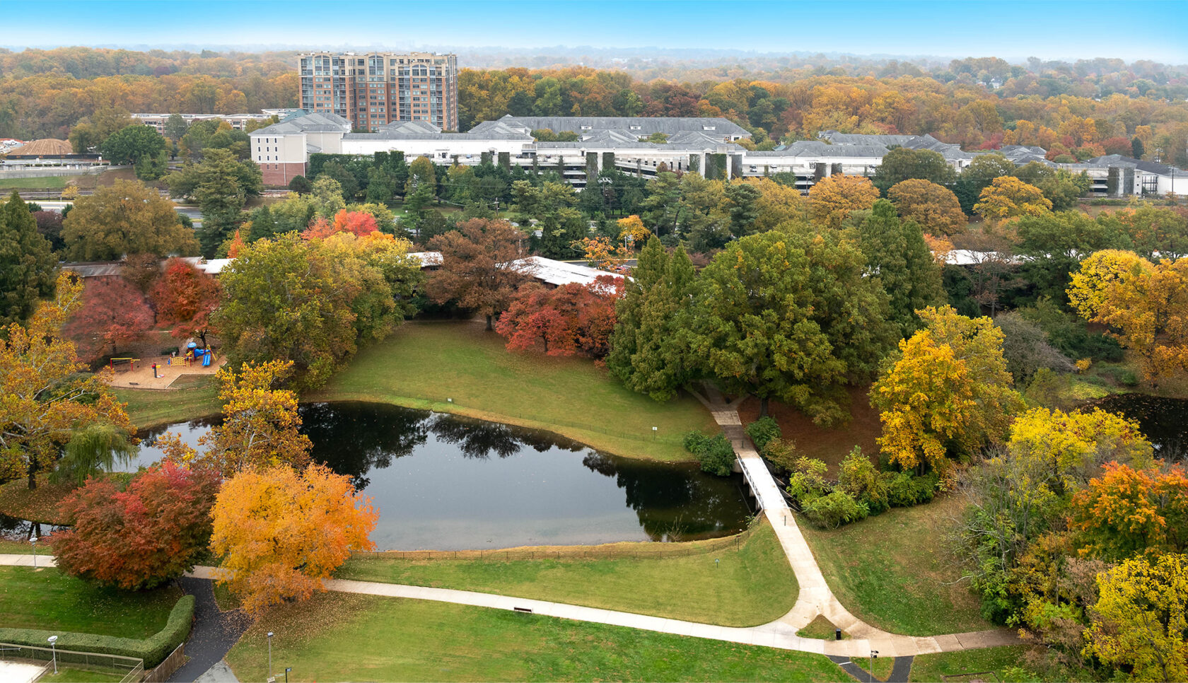 An aerial view of the back of the building, complete with walking paths, a pond, a playground and lots of trees.